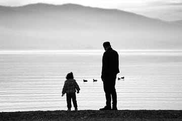 Silhouette of a man and child by a peaceful lake with ducks, black and white photo.